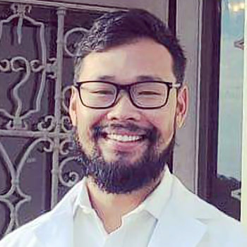 Man with glasses and a beard smiling, wearing a white shirt and jacket, standing in front of an ornate metal railing.