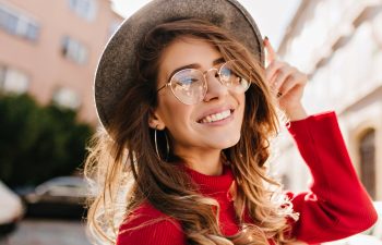 Woman in a red sweater smiles while touching her hat. She is wearing round glasses and hoop earrings, standing outdoors with buildings in the background.